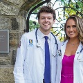 Twomedicalstudentswearingwhitecoatsandstethoscopessmilewhilestandingtogetheroutsidenearastonearchway