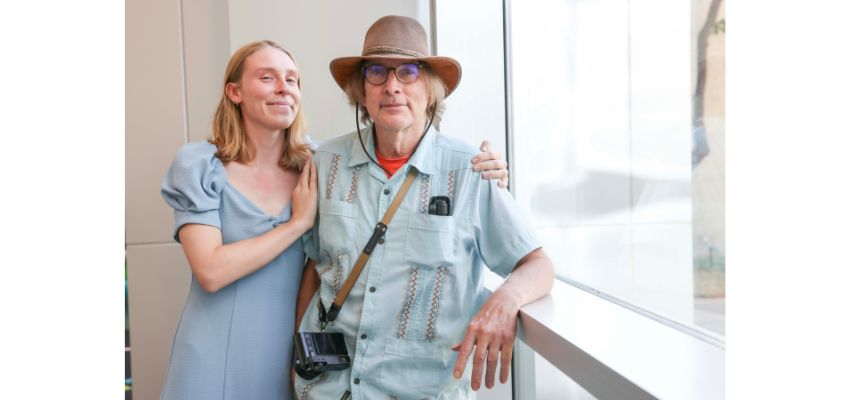 A woman in a light blue dress poses with an older man wearing glasses, a hat, and a camera strap inside a building with large windows.
