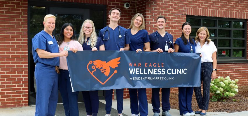 A group of students in scrubs and casual clothes stand in front of a brick building holding a banner that reads “War Eagle Wellness Clinic, A Student-Run Free Clinic.”