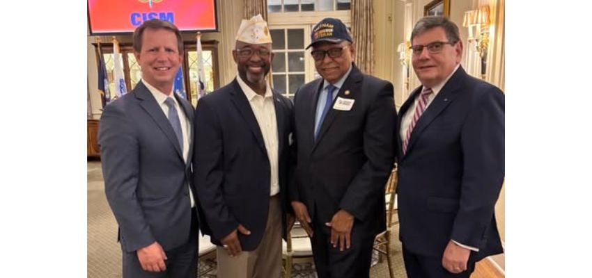Four men, including two in veterans’ hats, stand together in suits at an indoor event with flags in the background.