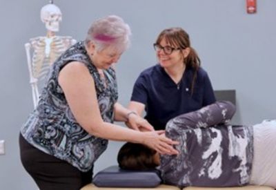A faculty member demonstrates a hands-on osteopathic technique on a patient while another student observes and smiles.