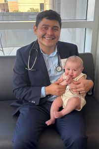 A medical student wearing a stethoscope and suit smiles while holding a baby on his lap.