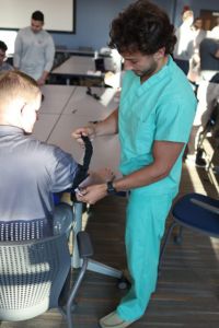 A student in scrubs places a blood pressure cuff on another student’s arm during a classroom activity.
