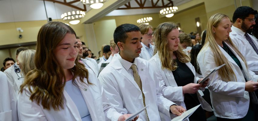Students wearing white coats stand in rows reading from booklets during a ceremony.