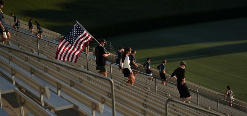 A group of people walk up stadium steps at sunrise, one carrying a large American flag.