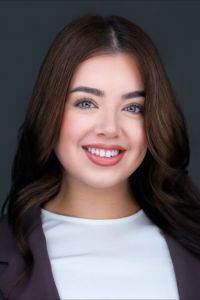 Headshot of a woman with long brown hair wearing a white top and dark blazer, smiling against a dark background.
