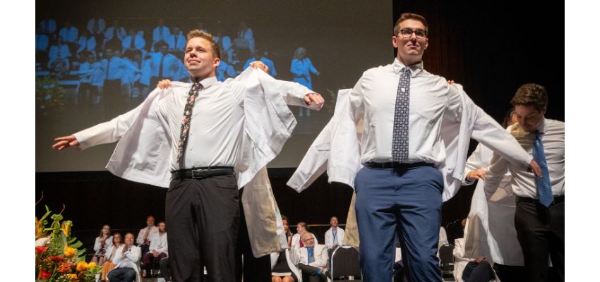 Students put on white coats on stage during a ceremony with an audience seated behind them.