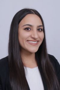 Headshot of a woman with long straight dark hair wearing a white top and black blazer, smiling against a light gray background.