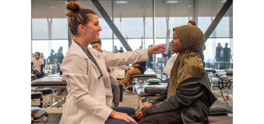A medical student in a white coat uses an eye chart to examine a young patient wearing a headscarf in a clinical training setting.