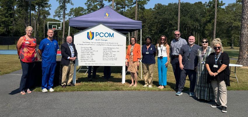 A group of people stand outside beside a PCOM South Georgia sign and tent, smiling in front of trees and a basketball court.