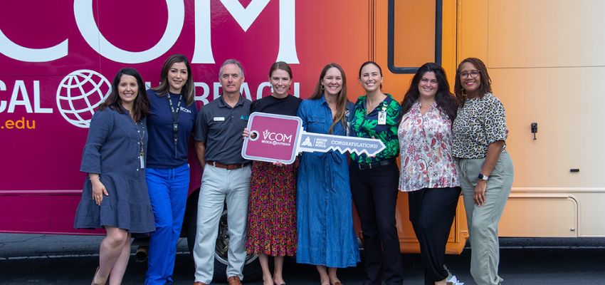 A group of people stand in front of a VCOM mobile medical outreach vehicle holding a large ceremonial key and smiling for the photo.