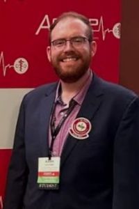 Aaron, wearing a blue blazer and conference badge, stands in front of the ACOFP step and repeat