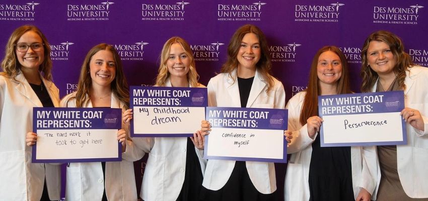 Six medical students in white coats stand in front of a Des Moines University backdrop, each holding signs sharing what their white coat represents.