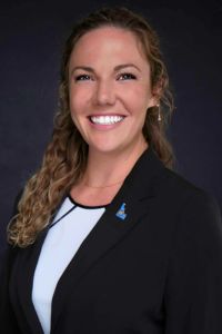 Headshot of a woman with curly hair wearing a black blazer and white blouse, smiling against a dark background.