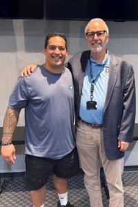 Two men stand side by side, one in a blue T-shirt and shorts and the other in a jacket and dress shirt, smiling with arms around each other.