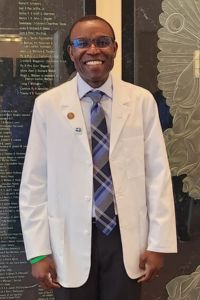 Man wearing a white coat and tie stands smiling in front of a wall with engraved names.