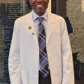 Manwearingawhitecoatandtiestandssmilinginfrontofawallwithengravednames