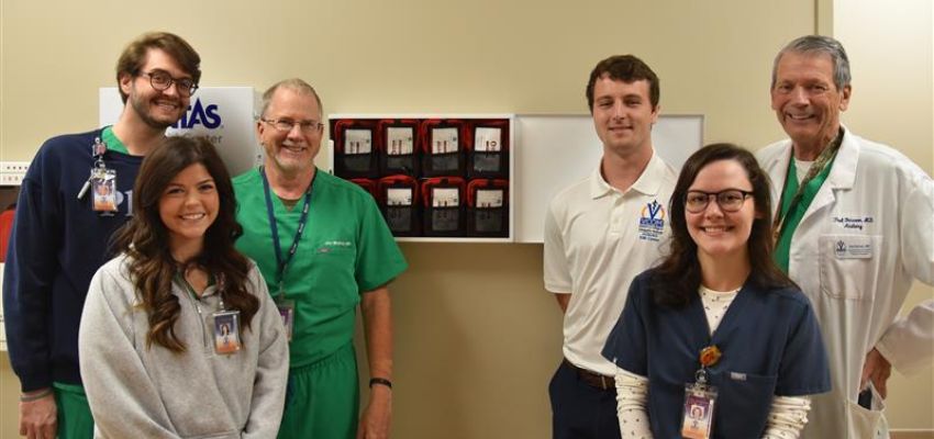 Six medical professionals and students smile for a group photo in a clinical setting, wearing scrubs and white coats.