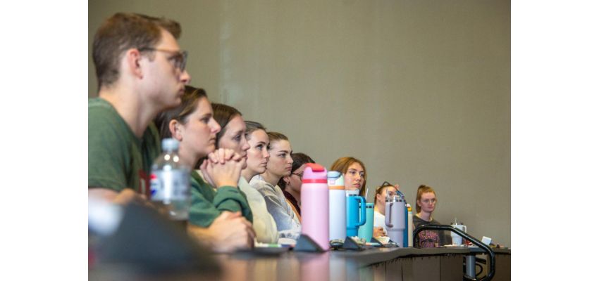 Students sit attentively in a lecture hall, focused on a presentation at the front of the room.