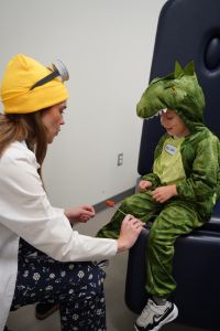 A medical student wearing a yellow hat and white coat interacts with a young child dressed in a green dinosaur costume during a playful clinical activity.