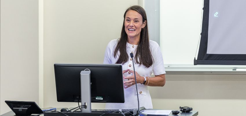 A woman in a white dress speaks at a podium in a classroom, smiling while giving a presentation.