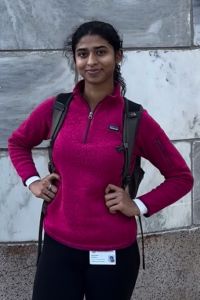 A student wearing a pink pullover and backpack stands confidently outside with hands on hips, smiling at the camera.