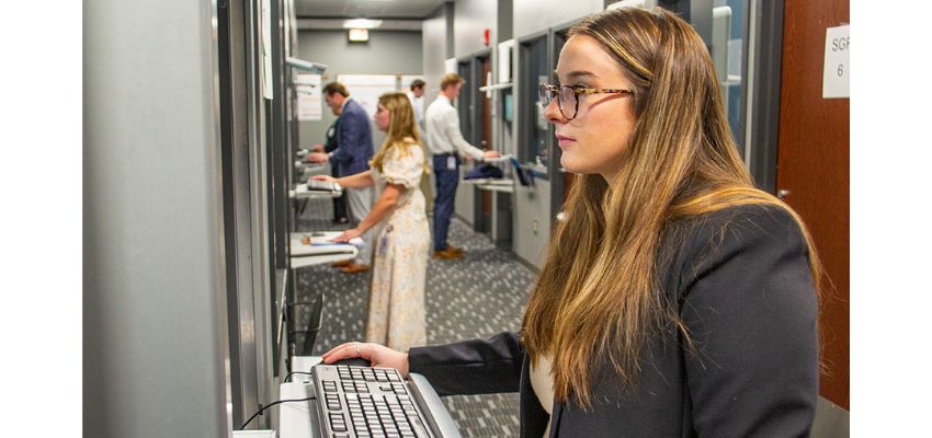 Students line up at computer stations in a hallway, taking part in an assessment or simulation activity.