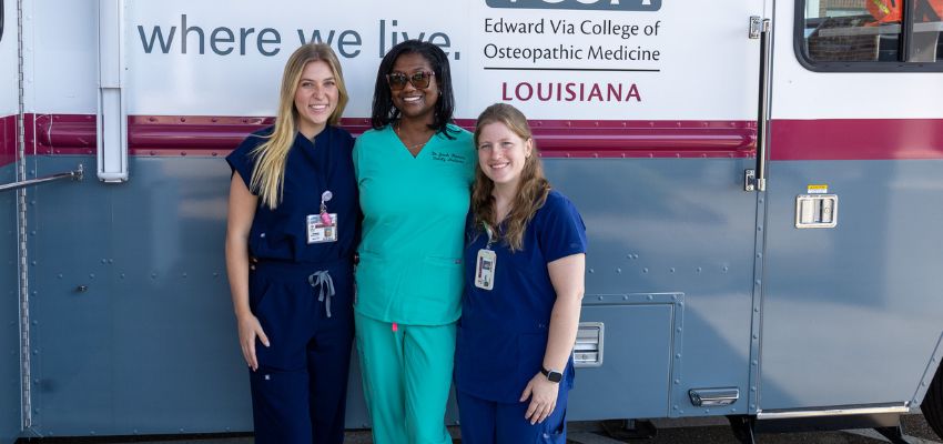 Three women in medical scrubs stand smiling in front of a VCOM Louisiana mobile clinic vehicle.