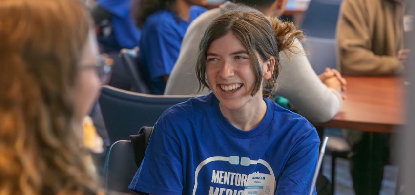 Student wearing a blue “Mentor Medics” shirt laughs while talking to another person during a mentorship event.