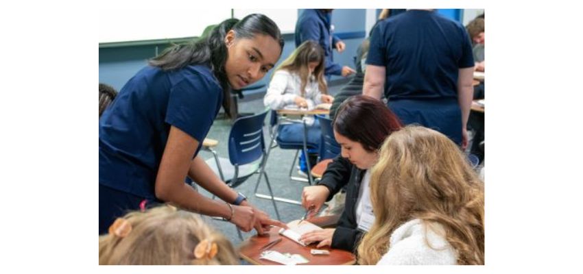 A student in scrubs assists a grade-school student with a hands-on medical skills activity in a classroom setting.