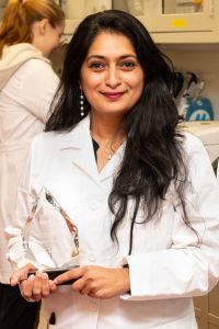 A woman in a whitecoat smiles while holding an award in a laboratory environment.