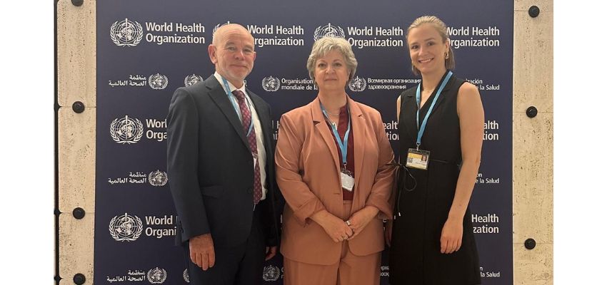 Three people stand smiling in front of a World Health Organization backdrop at an official event.