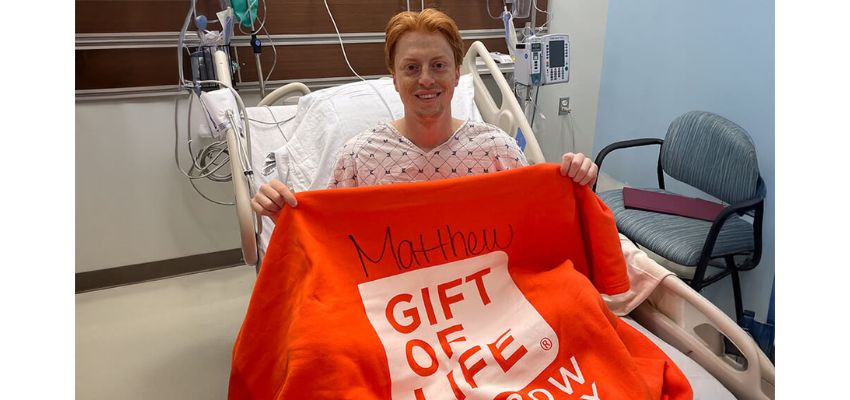 Matthew sits in a hospital bed after providing bone marrow, holding up a bright orange 