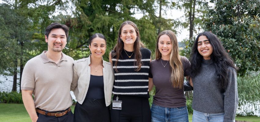 Five students stand together outside, smiling with trees and water in the background.