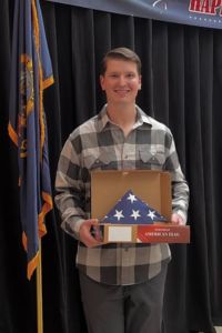 A young man stands indoors holding a folded American flag in a display box and smiling at the camera.