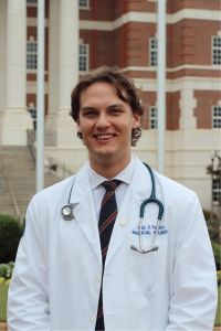 A medical student in a white coat stands smiling in front of a large brick building with columns.