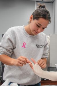 A student wearing a VCOM sweatshirt practices a clinical skill by working with another person's hand in a classroom setting.