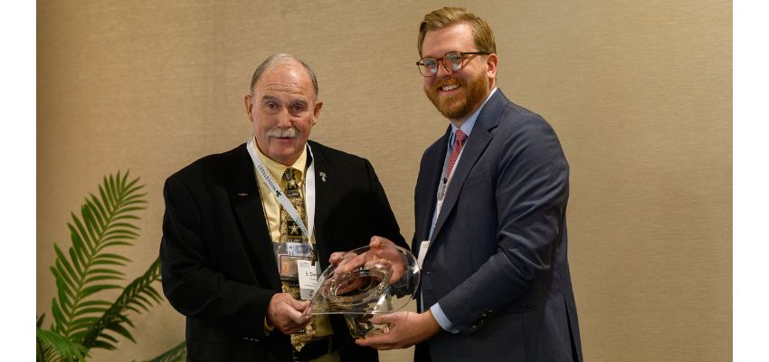A man in a suit presents an award to another man during an event, both smiling as they hold the glass trophy together.