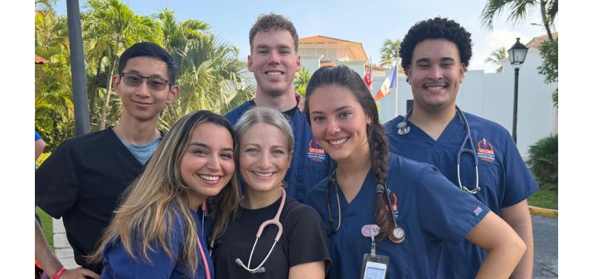 A group of smiling medical students in scrubs pose together outdoors with stethoscopes around their necks.