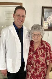 A medical student in a white coat stands smiling next to an older woman inside a home.