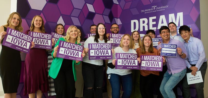 A group of medical students hold “I Matched in Iowa” signs while smiling together in front of a Des Moines University backdrop.
