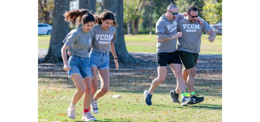 Students wearing VCOM shirts participate in a team-based outdoor activity on a grassy field.