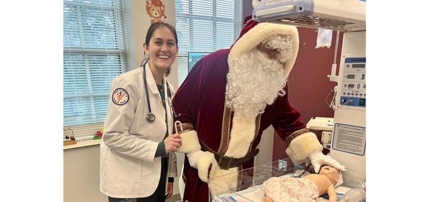 A medical student smiles beside a person dressed as Santa Claus while they care for an infant mannequin in a clinical setting.