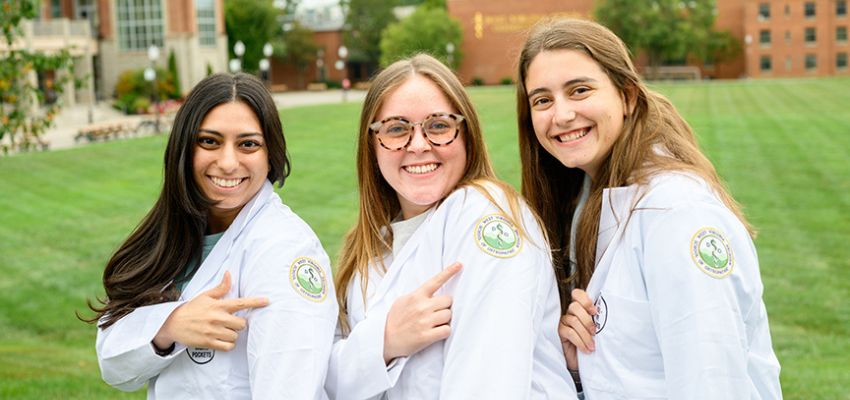Three medical students wearing white coats smile and point to a patch on their sleeves while standing on a campus lawn.