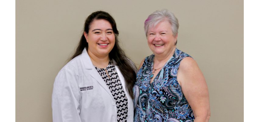 A woman in a white coat stands next to another woman in a patterned blouse, both smiling at the camera.