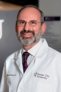 Dr. Hahn smiles in a professional environment, wearing his whitecoat with Kansas City University embroidered on it.
