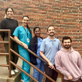 Agroupoffiveindividualsincludingmedicalprofessionalsinscrubsposetogetheronastaircaseoutsideabrickbuilding
