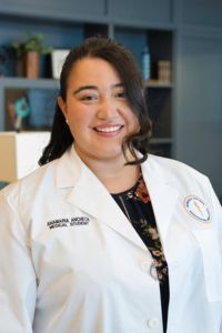A woman in a white coat stands indoors, smiling at the camera with shelves in the background.