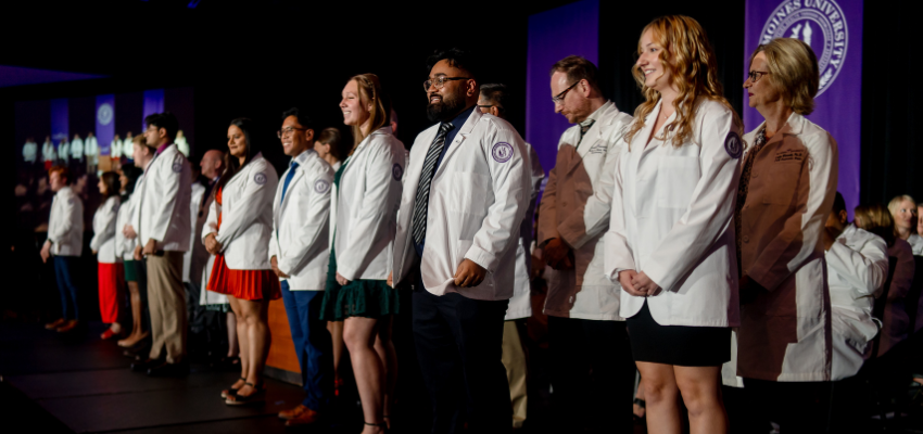 Students stand on stage while receiving white coats