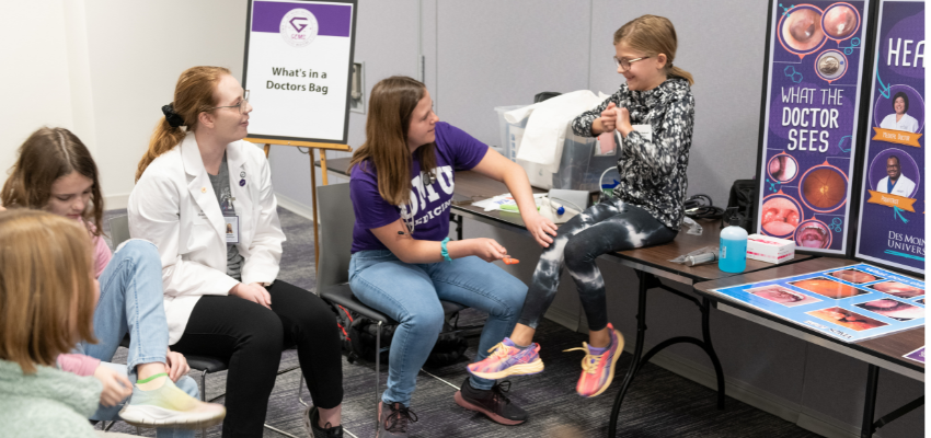 DMUCOM student checks reflexes on little girl sitting on table